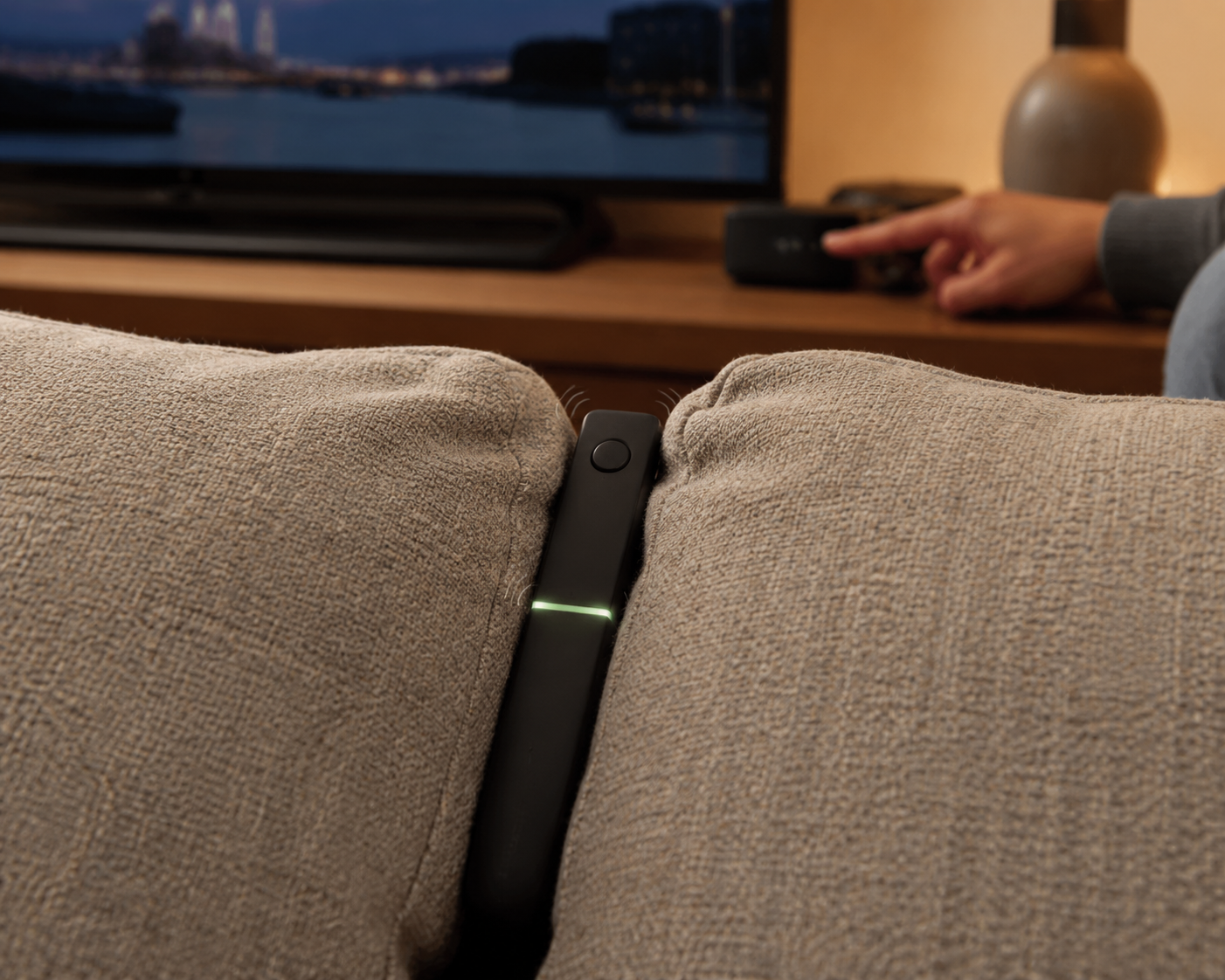 Concept visual of the remote lost between sofa cushions, flashing a green marker light while a hand presses the find button on a base unit beneath the television